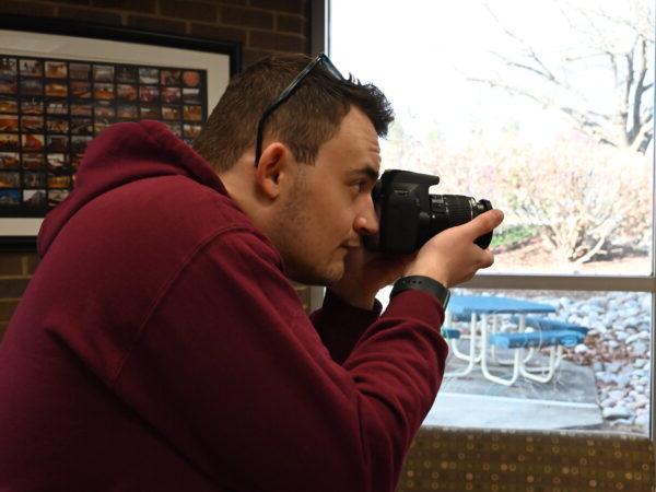 Image shows Brody Owens holding a camera up to his face, taking a photo in the Rothrock Library in front of a window.